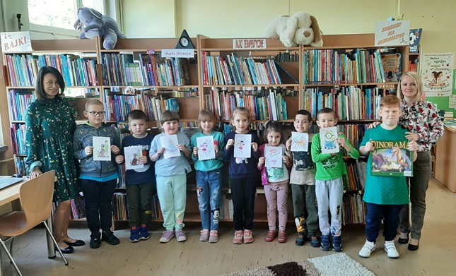 Grupa dzieci na tle książek pokazuje wykonane przez siebie kolorowanki.